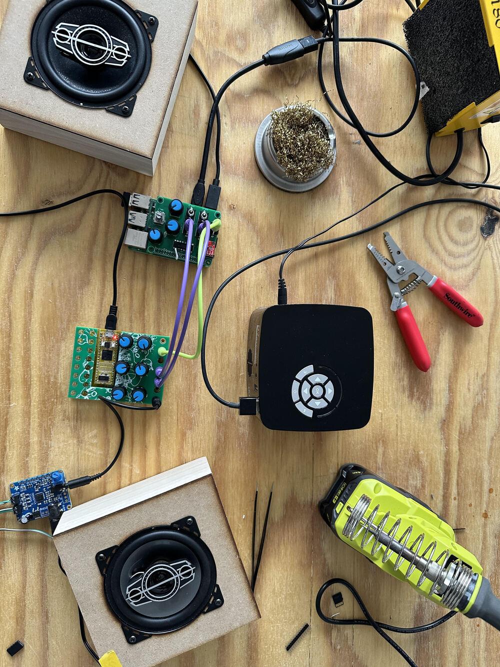 Overhead shot of Rpi with hat, with plug config described above. This time we see it runs to an EARTH synthesizer, a CCAM prototype. This sends audio out to a little amp board with DIY speakers. A small project is seen on the table top, as well as soldering supplies.
